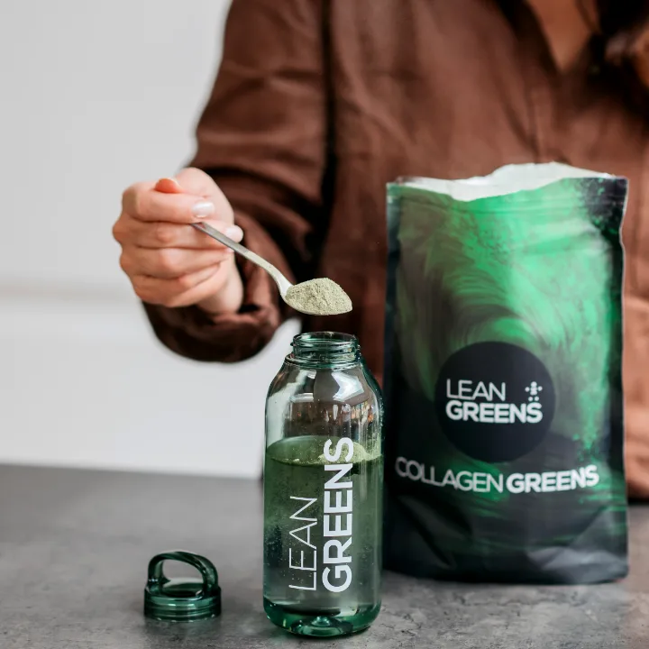 A teaspoon of Collagen Greens powder being tipped into a bottle of water beside the open pouch