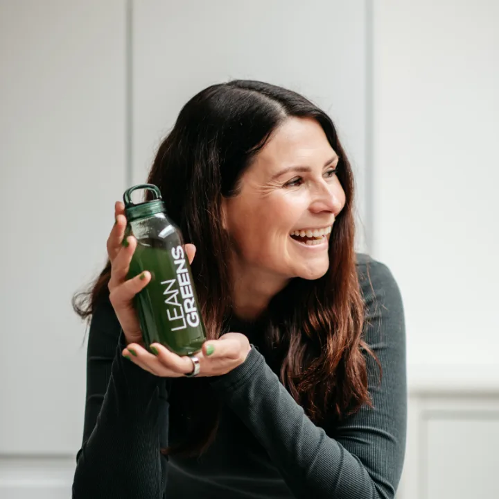 A woman laughing at her kitchen counter, holding a bottle of Collagen Greens